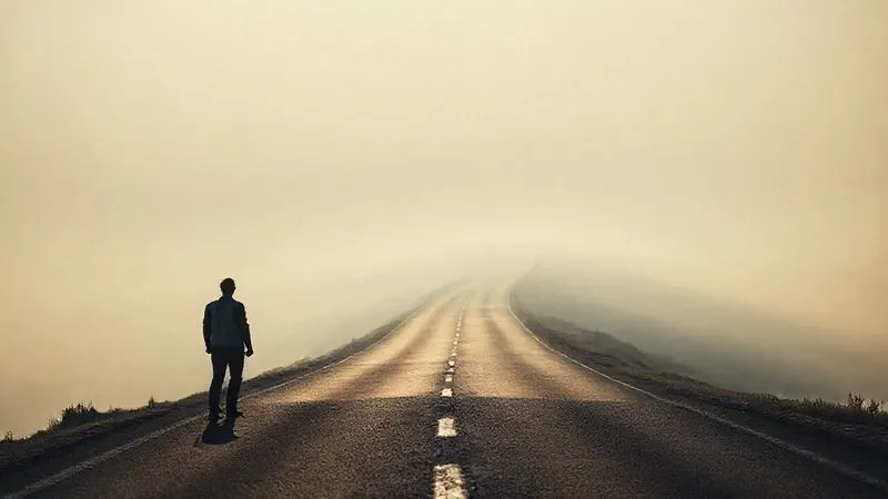 A photorealistic image of a lone figure standing at the base of a long, winding, and dimly lit road stretching into a hazy, uncertain horizon. The figure appears to be looking towards the distant future with a sense of apprehension. Cinematic lighting with subtle shadows, sharp focus on the figure and the immediate road, depth of field blurring the distant horizon. 8K hyper-detailed, shot on a high-end DSLR. Professional photography, emotionally resonant.